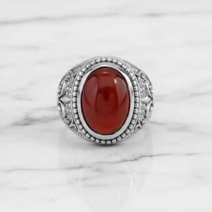 A silver-toned ring featuring an oval cabochon red gemstone, set with small clear stones in a decorative border, displayed on a white marble surface.