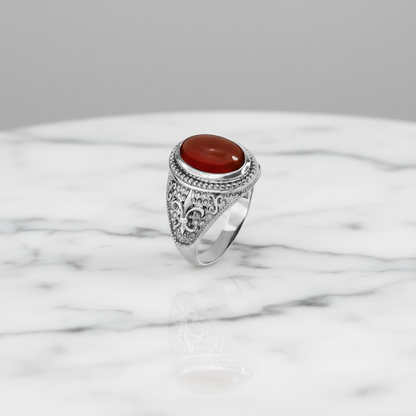An ornate sterling silver ring with a round red cabochon gemstone, displayed on a white marble surface against a neutral gray background.