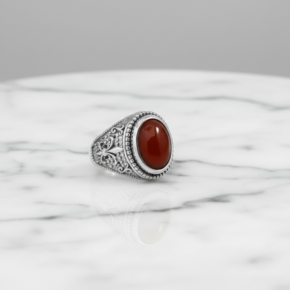 A silver signet ring with an oval red cabochon stone, displayed on a white marble surface against a neutral gray background.