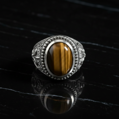 A silver-toned men's ring featuring an oval cabochon tiger's eye stone with bold brown and gold stripes, accented by small clear stones on the shoulders, displayed on a glossy black reflective surface.