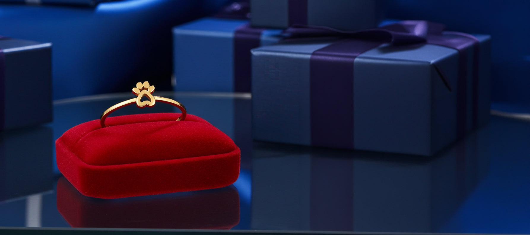 A gold ring with a small paw-print clasp displayed on a red velvet ring box, with blue gift boxes in the background.